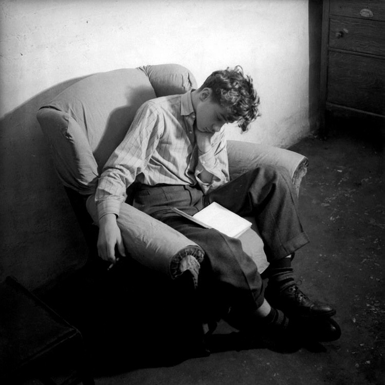 a black and white photo of richard soakup - a young white man with short curly brown hair - sitting in an armchair with his head rested on his left hand, seemingly asleep, with an open book in his lap and cigarette in his right hand that is hanging down from the armchair. he is dressed in a white pinstriped button up shirt with 2 buttons undone, grey pleated dress pants, warm socks and black dress shoes